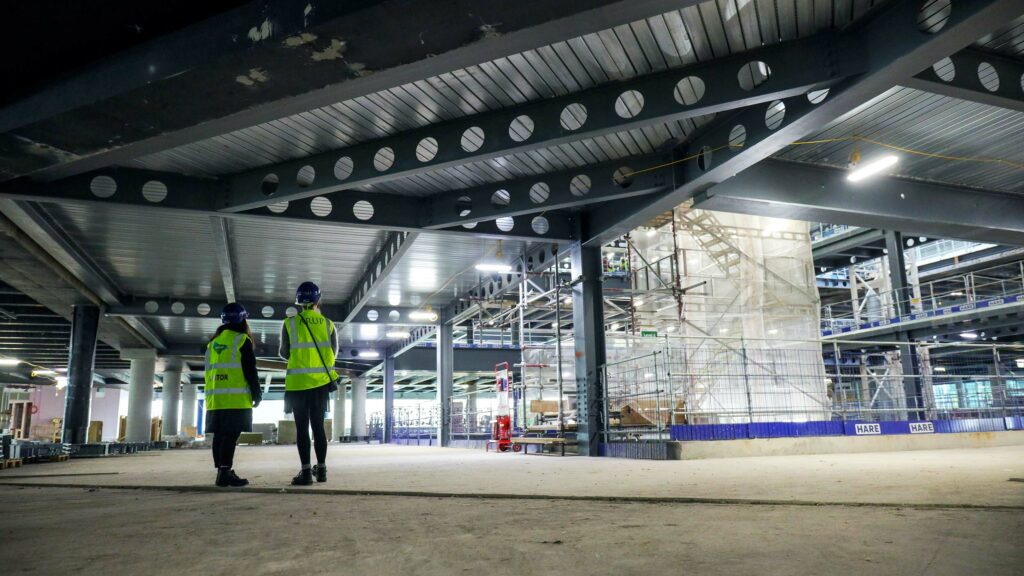 A photograph of the inside of a large construction site. We can see the backs of two women wearing PPE.