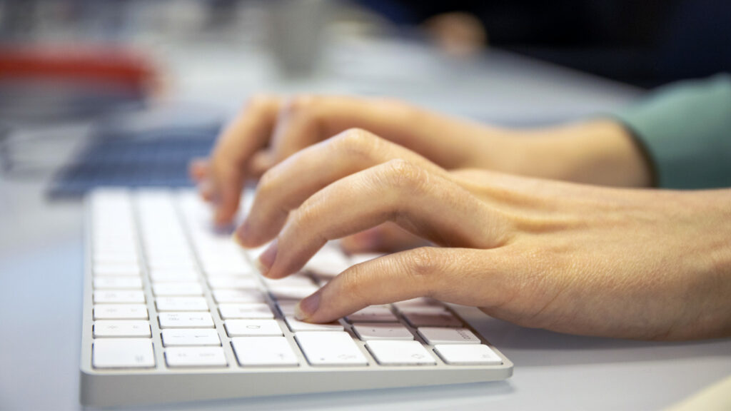 A close up photograph of hands typing at a keyboard.